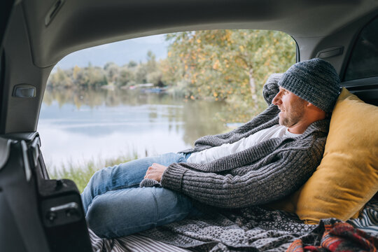 Smiling middle-aged man dressed in warm knitted clothes and jeans lying in the cozy car trunk and enjoying the mountain lake view. Warm early autumn auto traveling concept image.