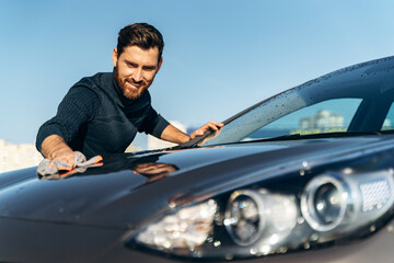Waist up portrait view of the young happy male owner cleaning his black car with cloth while...