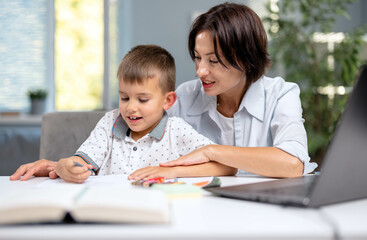 Loving caucasian mother sitting with her son at desk and helping with homework. Cute boy writing and reading at home. Domestic education and parenting concept.