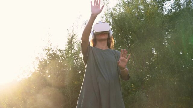 A girl with virtual reality glasses on her head, standing against the background of green trees. He moves his hands through the air. The concept of the future. High quality 4k footage