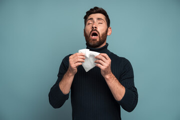 Coughing or sneezing. Sick man sneezing tissue, coughing, feeling unwell, sick or flu, suffering allergy or seasonal influenza symptoms. Indoor studio shot isolated on blue background