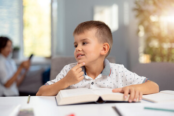 Cute little boy sitting at desk with books and notes and dreaming about something. Caucasian schoolboy looking aside during domestic education.
