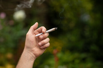 Mans hand holding a weed joint in a green surrounding with nature, plants and greenery. Smoking...