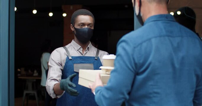 Close up. Rear of old Caucasian customer take out food bag order with coffee from restaurant staff waiter wearing face mask and gloves in coffee shop. New normal, coronavirus take away food service