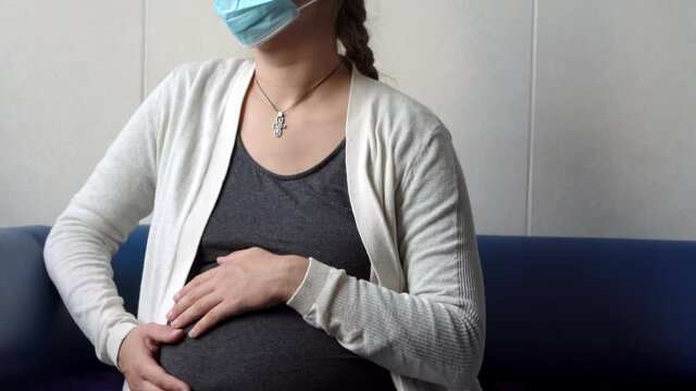 Young Pregnant Woman In Medicine Protective Mask Reading On Couch Waiting For Appointment Medical Control On Hospital. Visit Gynecologist Doctor At Clinic. Examine Expectant Belly Healthcare Check Up