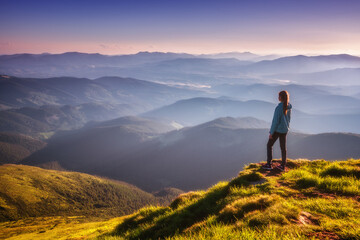 Girl on mountain peak with green grass looking in beautiful mountain valley in fog at sunset in autumn. Landscape with sporty young woman, foggy hills, forest, sky in fall. Travel and tourism. Hiking - obrazy, fototapety, plakaty
