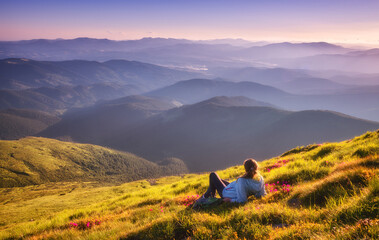 Young woman on the mountain peak with green grass and pink flowers looking at beautiful mountains...