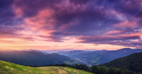 Fototapeta na wymiar Mountains at beautiful sunset in summer. Colorful panoramic landscape with meadows with green grass, sky with vibrant clouds, mountains with forest. Trail on the hill. Travel and nature. Scenery