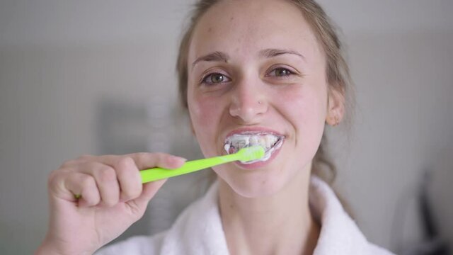 Close-up of brunette young woman with brown eyes brushing teeth in slow motion smiling looking at camera. Portrait of confident slim beautiful Caucasian lady taking care of dental health at home