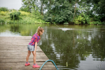A blonde girl stands on a wooden pier on the river bank.