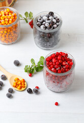 Frozen berries of cranberries, blueberries or blueberries and sea buckthorn in glass jars. On a light gray background, vertical