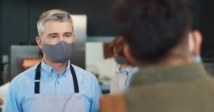 Close up of cheerful handsome Caucasian middle-aged grey-haired male barista in mask standing at counter in own coffee house talking to man client. Take away food and drink service, business, covid-19