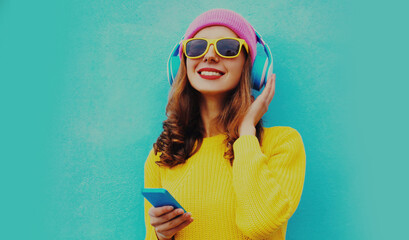 Portrait of modern young woman in wireless headphones listening to music with phone wearing yellow knitted sweater and pink hat on blue background