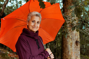 portrait of mature woman with gray hair looking at camera holding an orange umbrella on a rainy...