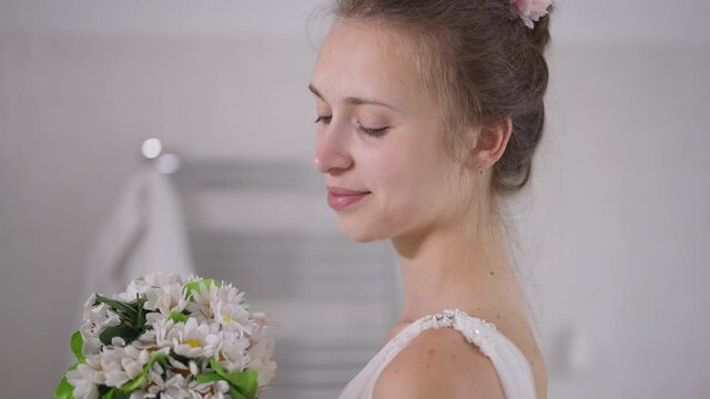 Side view close-up portrait of happy charming young Caucasian bride admiring white bridal bouquet turning looking at camera smiling. Confident beautiful slim woman posing indoors in the morning