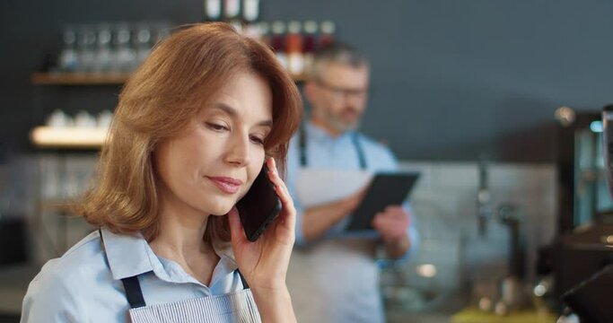 Close up portrait of beautiful happy woman in apron speaking on smartphone at workplace in cafe while male colleague typing on tablet on background. Family business, coffee house, job concept