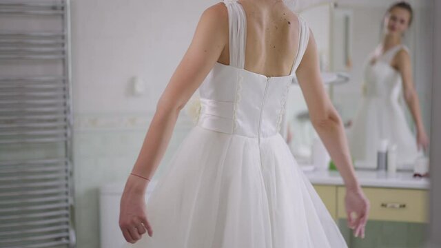 Unrecognizable slim happy young bride spinning in slow motion in white wedding dress with blurred reflection in mirror at background. Joyful excited Caucasian slim woman enjoying morning