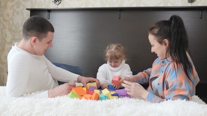 Obraz na płótnie Canvas Parents, dad, mom, daughter play with cubes in nursery on bed. Teaching child through play. Child, father and mother are playing building family home. Happy family. Educational games for children.