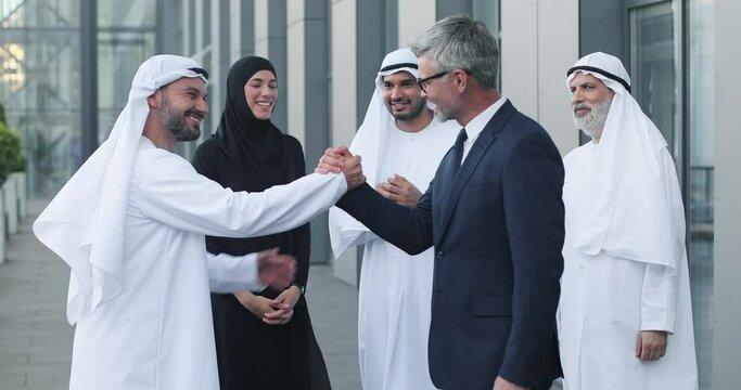 Nice to meet you. Portrait of cheerful muslim entrepreneur wearing kandura standing at the street and having firm handshake with new employee. Their colleagues expressing gladness and applauding