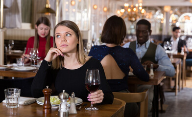 Pensive young woman sitting alone in restaurant table with glass of red wine