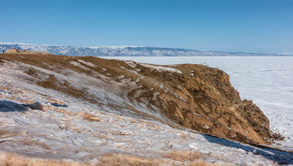 The frozen lake has steep shores. The snow lies on the bare ground and on the ice. Tiny silhouettes of people walking are visible. A mountain range against the blue sky. Baikal