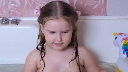 Little playing girl 3 years old with two ponytails on her head, bathes and washes in the water in the bathroom at home. Baby body care concept, hygiene.