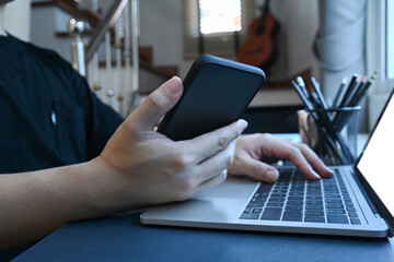 Close up man holding mobile phone and working with computer laptop at home.
