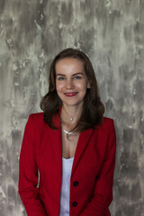 Portrait of a beautiful young business woman with brown hair in a red jacket and white blouse. Happy smiling girl with red lips on gray background with copy space. The concept of business and success.