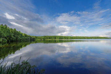 Sunny day on a calm river in summer. The blue sky is reflected in the water. Landscape with a lake and a strip of forest on the horizon.