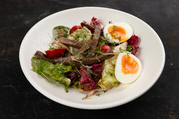A beautiful Fresh salad with meat tongue vegetables lettuce leaves egg cucumbers bell pepper Cherry tomatoes olives red onions is decorated on a white plate on a black background large serving 