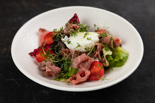 A beautiful Fresh salad with meat roast beef vegetables lettuce leaves poached egg cucumbers bell pepper olives red onions is decorated on a white plate on a black background large serving Restaurant