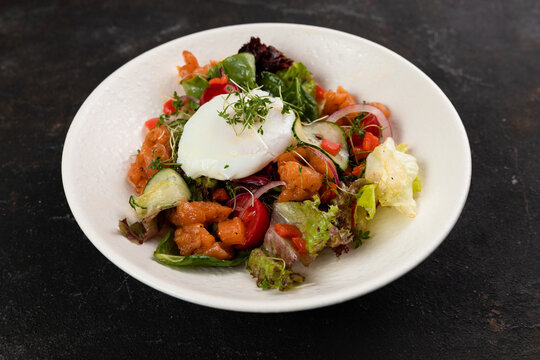 A beautiful fresh salad with salmon red fish vegetables lettuce leaves poached egg cucumbers bell peppers olives red onions is decorated on a white plate on a black background close up