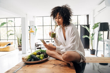 young mixed race woman looking at cellphone having green juice breakfast. sitting on kitchen...