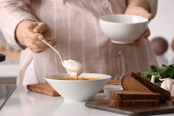 Woman adding sour cream to bowl with delicious borscht in kitchen