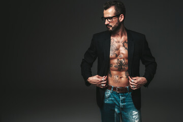 Obraz premium Portrait of handsome confident stylish hipster lambersexual model. Sexy modern man dressed in black jacket. Naked torso with tattoos.Fashion male posing in studio on dark background. In spectacles