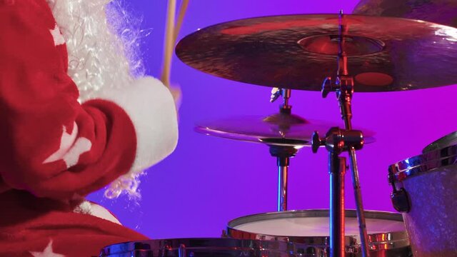 Side view of Santa Claus in a red costume with beard plays the drums in a dark studio with purple lights. A man in a carnival costume performs a show at a New Year's party. Close up. Slow motion.
