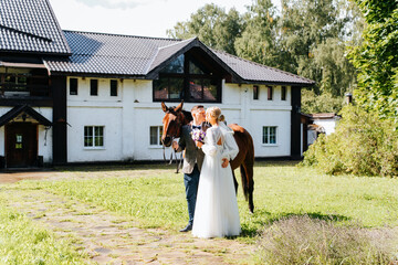 Beautiful young married couple with horse at villa, newlywed wedding kiss at ranch outdoors. Bride...