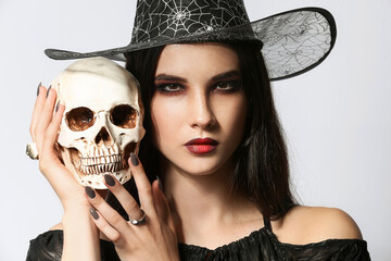 Young witch with human skull on light background