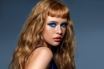 An attractive beautiful woman with natural curly shiny hair poses. A lot of hairstyle with the effect of beach curls. Red-haired high fashion model with freckles on her face with blue evening make-up.