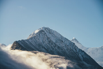 Fototapeta premium Beautiful view of snow-capped mountains above thick clouds in sunshine. Scenic bright mountain landscape with white-snow peak among dense low clouds in blue sky. Wonderful scenery with snowy pinnacle.