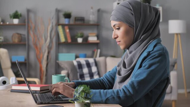 Focused Muslim Woman Wearing Hijab Typing At The Laptop Computer Working From Home,side View Of Concentrated Arab Female Student Sitting At Her Desk Using Notebook