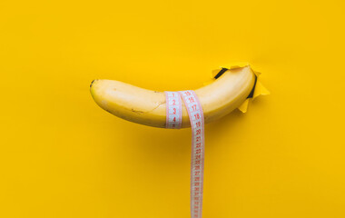A large banana with a tape measure wrapped around it appears through a torn hole in yellow paper....
