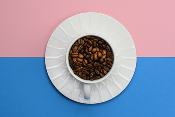 White cup and saucer filled with roasted coffee beans on a blue and pink background. Flat lay, top view.