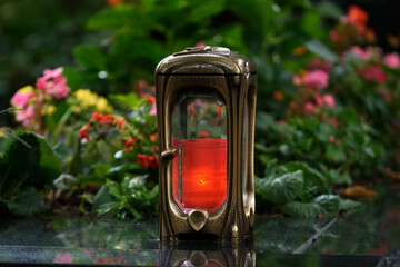 metal grave lamp with burning candle on a grave with colorful  flowers in blurred background