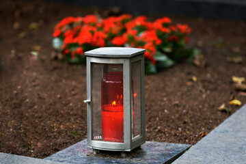 metal grave lamp with burning candle on a grave with red flowers in blurred background