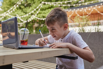little schoolboy doing homework outdoors with laptop. smiling caucasian boy sitting at table at veranda of cafe, studying with computer, writing notes in copybook. distant learning, online education