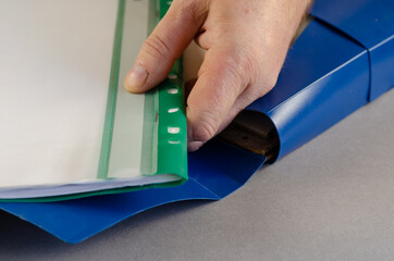 A mature male hand is holding a green file folder. Blue document case on gray background. Open briefcase with reports, technical documentation. Close-up. Selective focus.