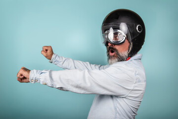 Profile of bearded hipster in denim shirt with helmet and glasses pretending to ride a motorcycle, isolated on blue studio wall.