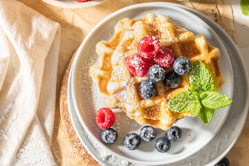 Traditional Belgian waffles stacked with fresh raspberries and blueberries sprinkled with powdered sugar on the kitchen counter