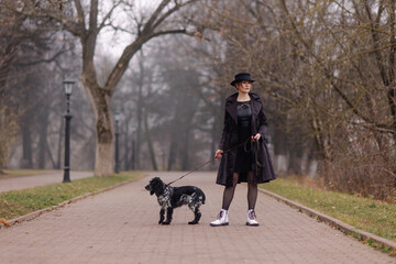 woman walks with a dog on a leash in the park in autumn. November October chilly weather fog. owner and pet in collar. puppy is the best friend. Homeless. Girl in a black cloak and hat. Mood of winter